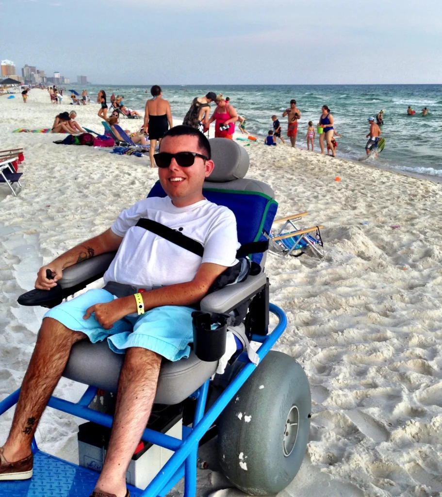 A person sits in a beach wheelchair on sandy shore, surrounded by beachgoers enjoying the sun and water in the background.