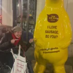A yellow bear statue with a crown, holding a sign that says "I LOVE SAUSAGE. DO YOU?" in front of a restaurant at night.