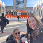 A vibrant outdoor event scene with attendees in formal attire, colorful flags, and a large sign reading "ITB Berlin" in the background.