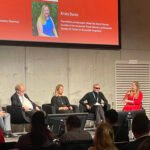 A panel discussion featuring four speakers, with a PowerPoint presentation displaying participants' names and titles in a conference setting.