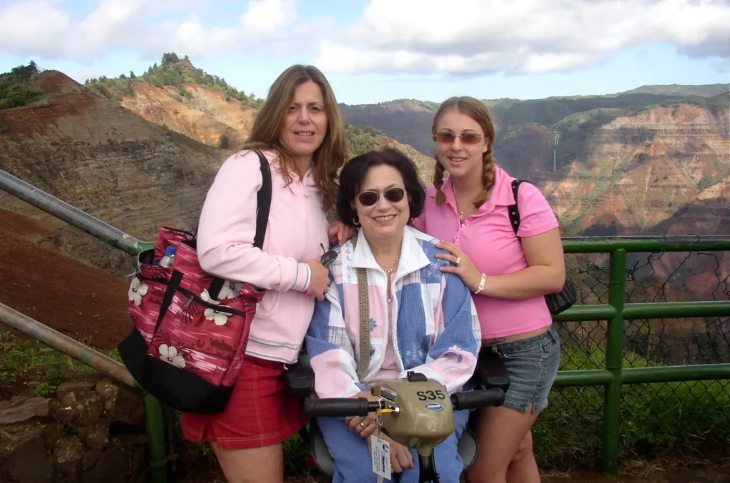 Three women pose together at a scenic overlook, with colorful hills and a partly cloudy sky in the background.