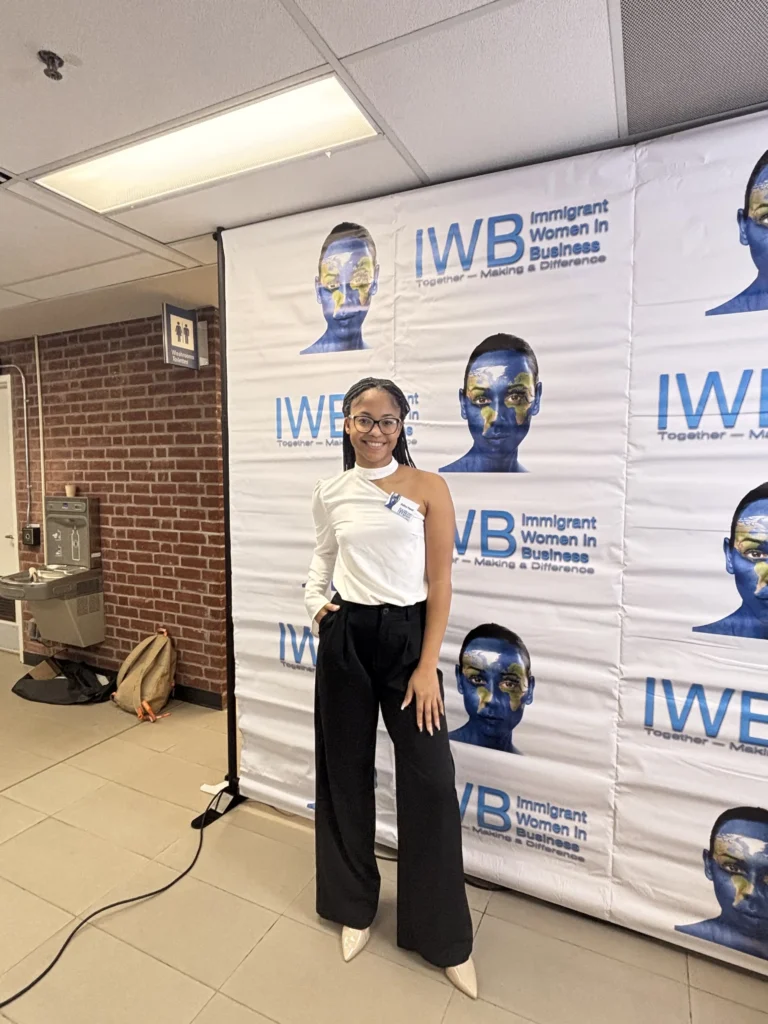 A woman poses confidently in front of an IWB banner, wearing a stylish outfit with a white top and black pants.
