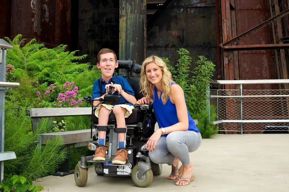 A woman kneeling next to a smiling man in a wheelchair, set against an industrial-style backdrop with greenery and wildflowers. Both are casually dressed and appear happy, depicting inclusivity and friendship.