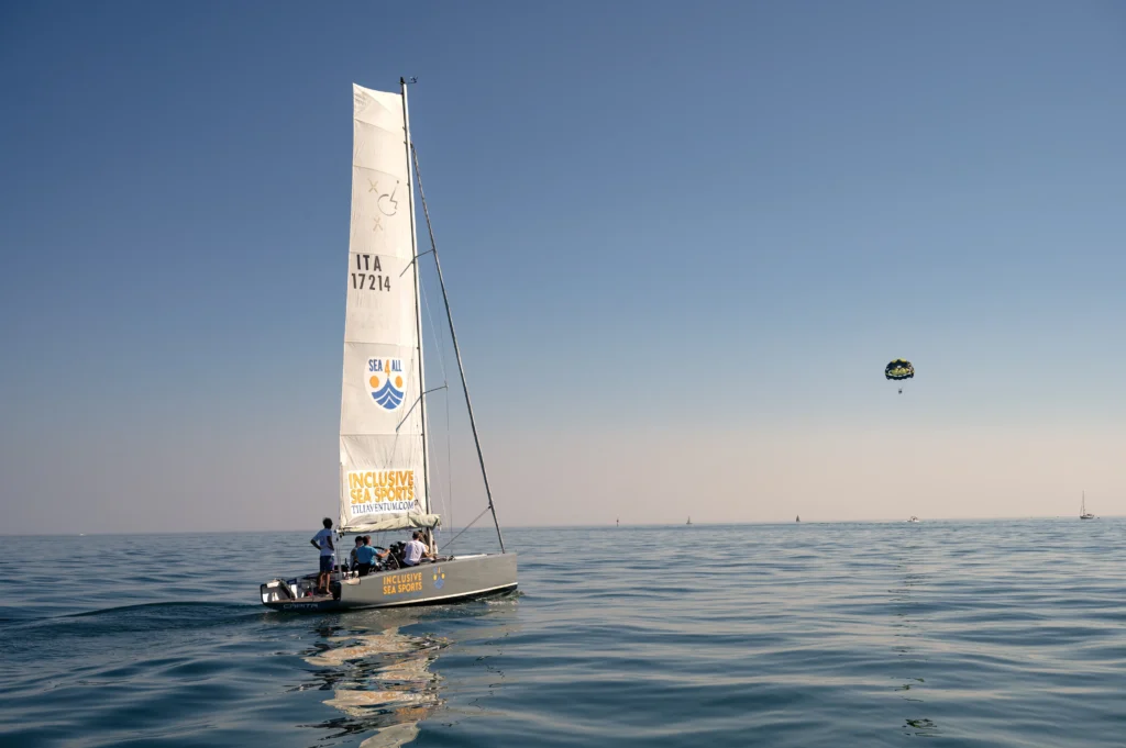 A sailboat with an inclusive sports logo sails on calm waters, while a person parasails above against a clear sky.
