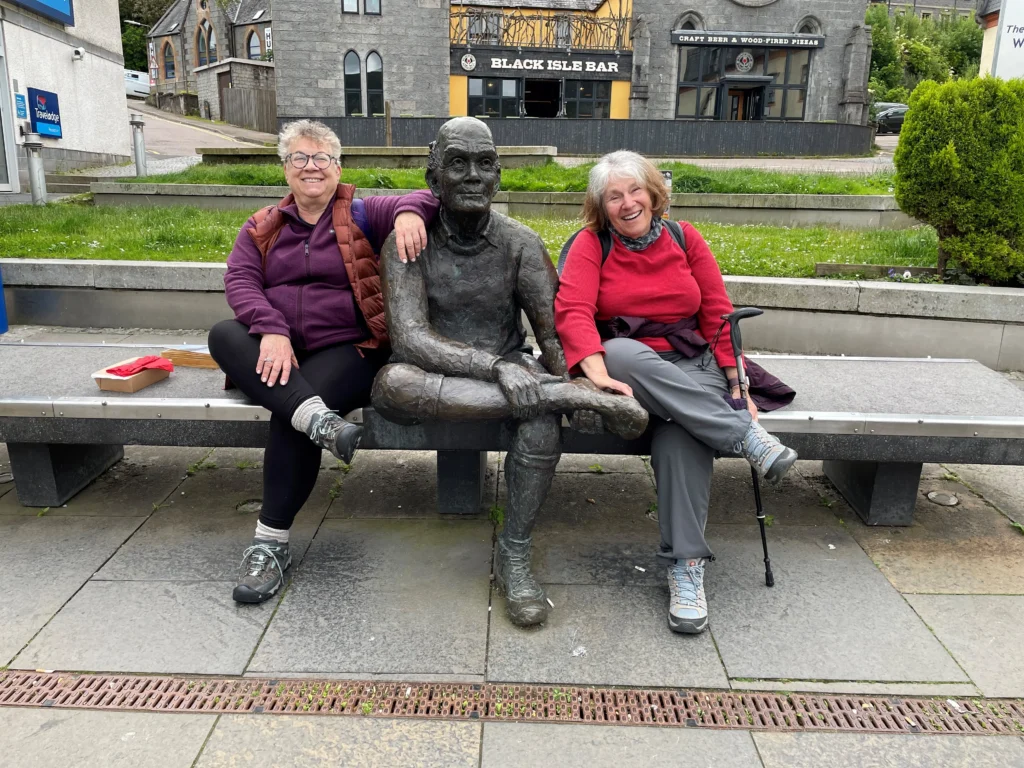 Two women sit on a bench beside a bronze statue, surrounded by green grass and a backdrop of buildings, enjoying their time together.