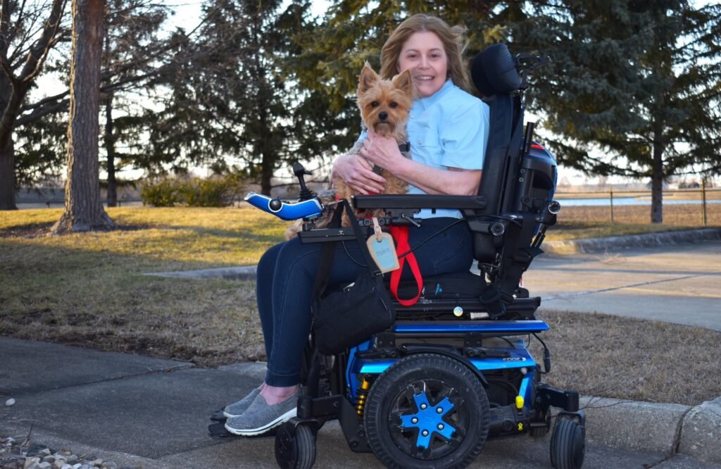 A woman sits in a motorized wheelchair on a paved sidewalk, smiling and holding a small brown dog in her arms. She is wearing a light blue shirt and dark jeans, and the dog appears calm and content. The wheelchair is black with blue accents and is equipped with various accessories, including a bag and a red lanyard with an ID card. In the background, there are leafless trees and a grassy area, suggesting a cool season.