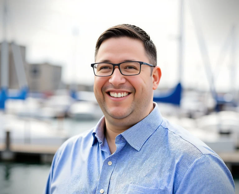A person wearing a light blue checkered shirt stands near a marina, with boats and masts visible in the background.