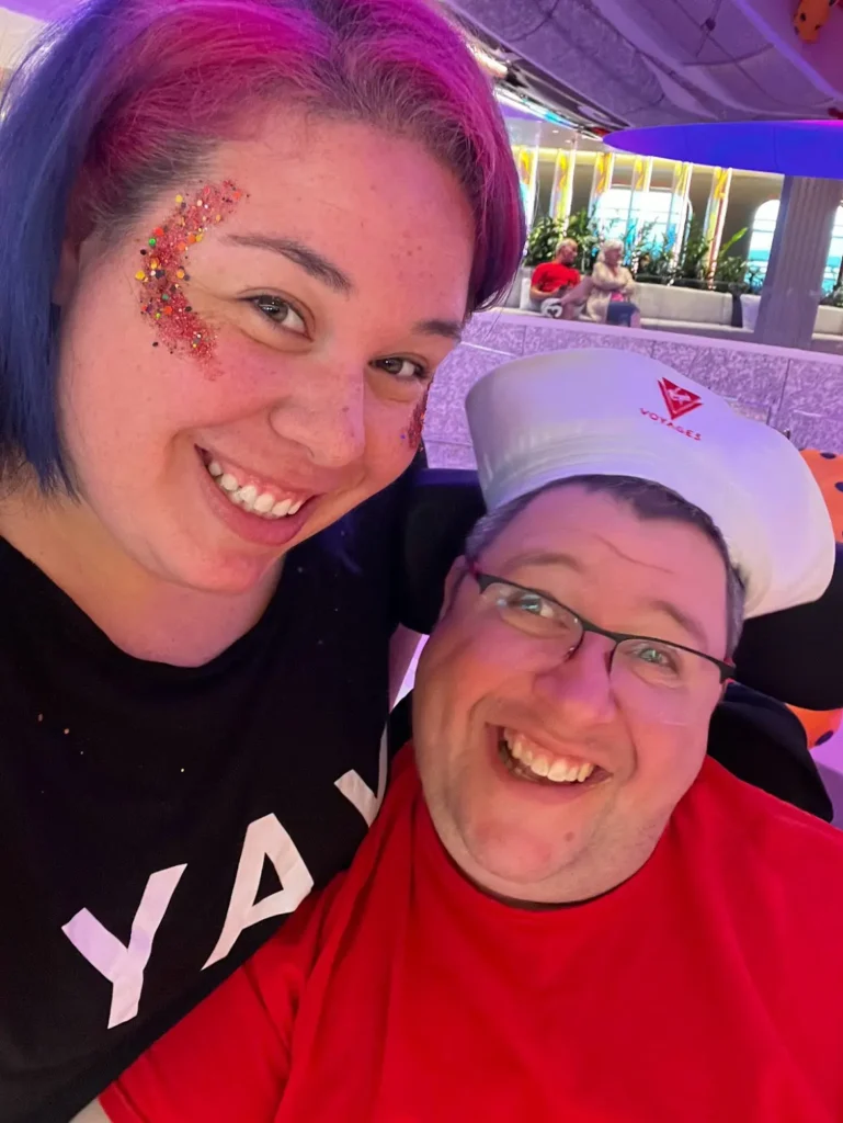 Two people pose for a selfie, one with colorful hair and a black "YAY" shirt, and the other in a red shirt, with a vibrant background.