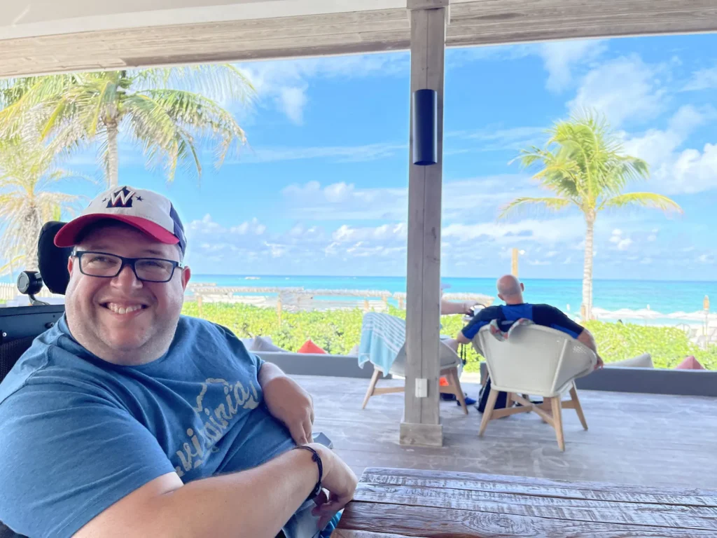 A man in a blue shirt and cap sits in a chair, overlooking a vibrant beach with palm trees and a clear blue sky in the background.