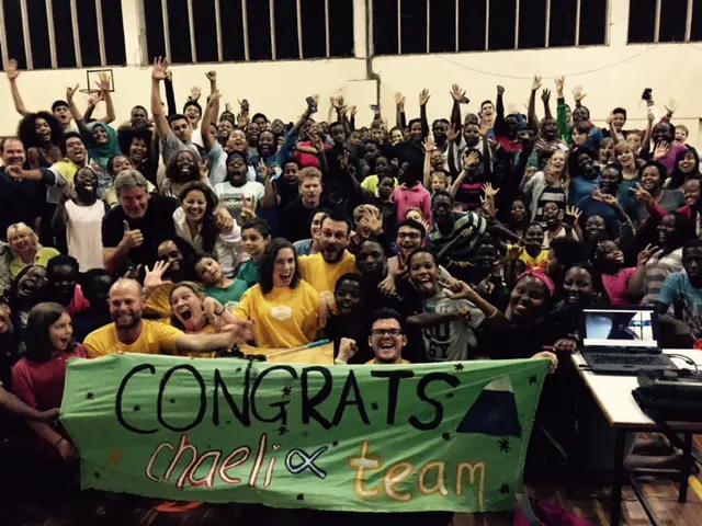 A large diverse group of people celebrating together indoors, holding a banner that reads "CONGRATS chaeli team" with joyous expressions.