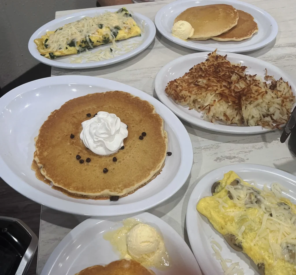 A colorful breakfast spread featuring fluffy pancakes with whipped cream, an omelet with cheese, and crispy hash browns on white plates.