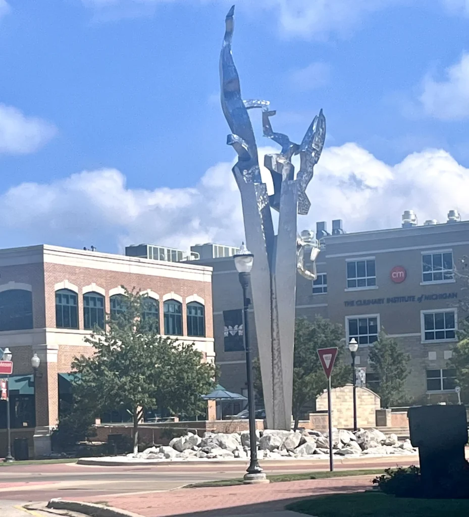 A tall, abstract silver sculpture rises above a landscaped area near a brick building, with a blue sky dotted with clouds.