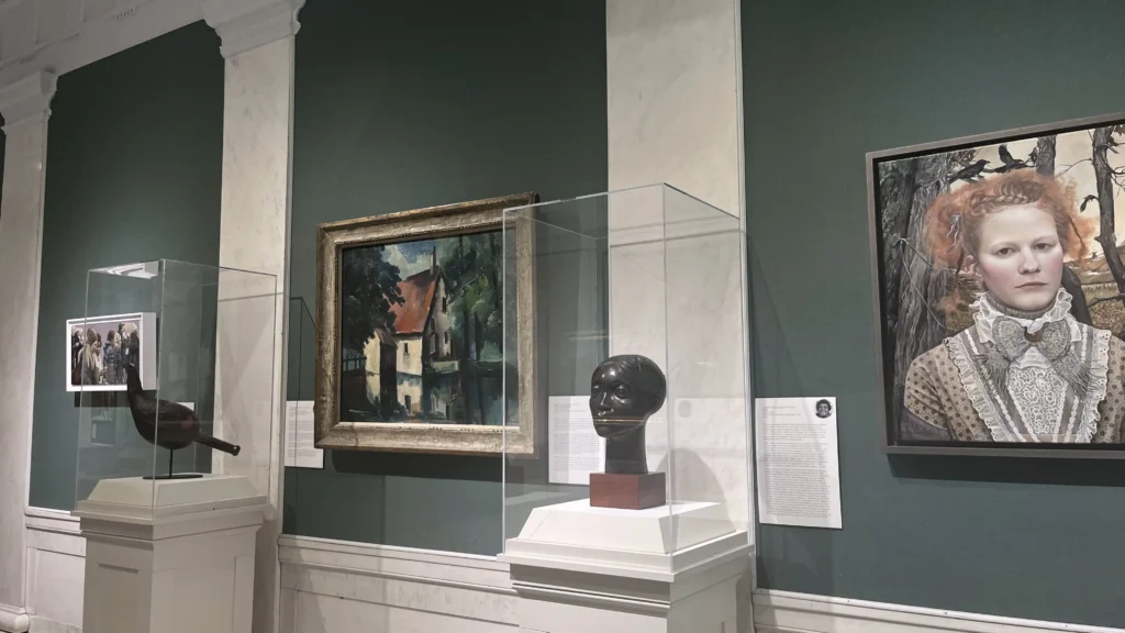 A museum interior featuring various artworks: a landscape painting, a displayed sculpture, and a framed photo of historical figures.