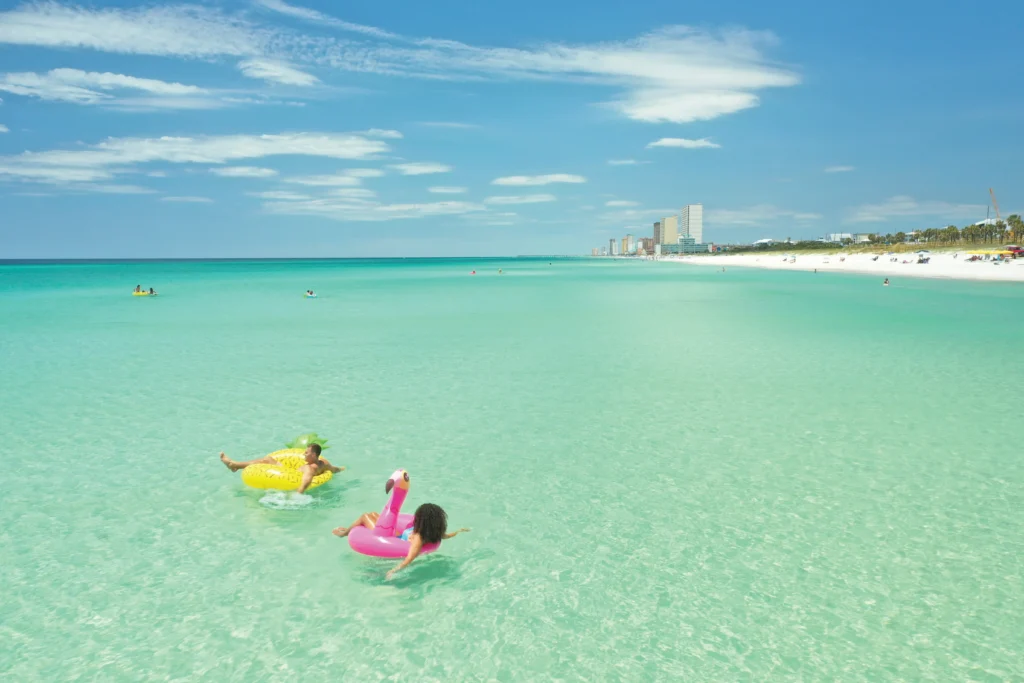 Two children float in clear turquoise water on inflatable rings, with a sandy beach and tall buildings in the background under a bright blue sky.