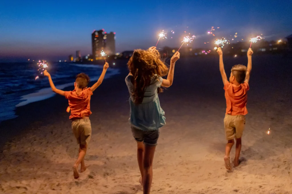 Three children on a beach at dusk joyfully wave sparklers, with city lights twinkling in the background and waves gently lapping at the shore.