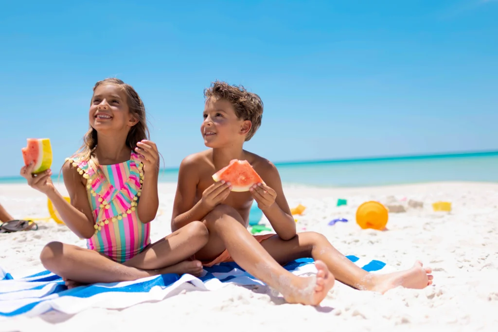 Two children sitting on a beach towel, enjoying slices of watermelon, with a clear blue sky and ocean in the background.