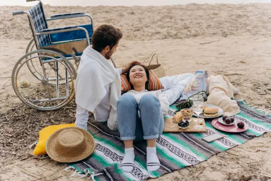 A couple enjoys a beach picnic on a colorful blanket with food and drinks. The woman lies smiling, leaning on the man's lap. A wheelchair is nearby.