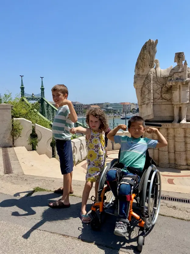 Three children pose confidently outdoors; one in a wheelchair, flexing their arms against a sunny backdrop with a historic statue and bridge.