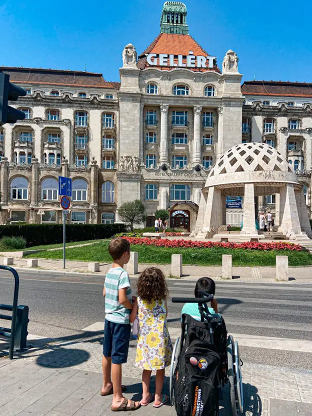 Three children, one in a wheelchair, stand in front of the Gellért Hotel in Budapest, admiring its grand architecture on a sunny day.