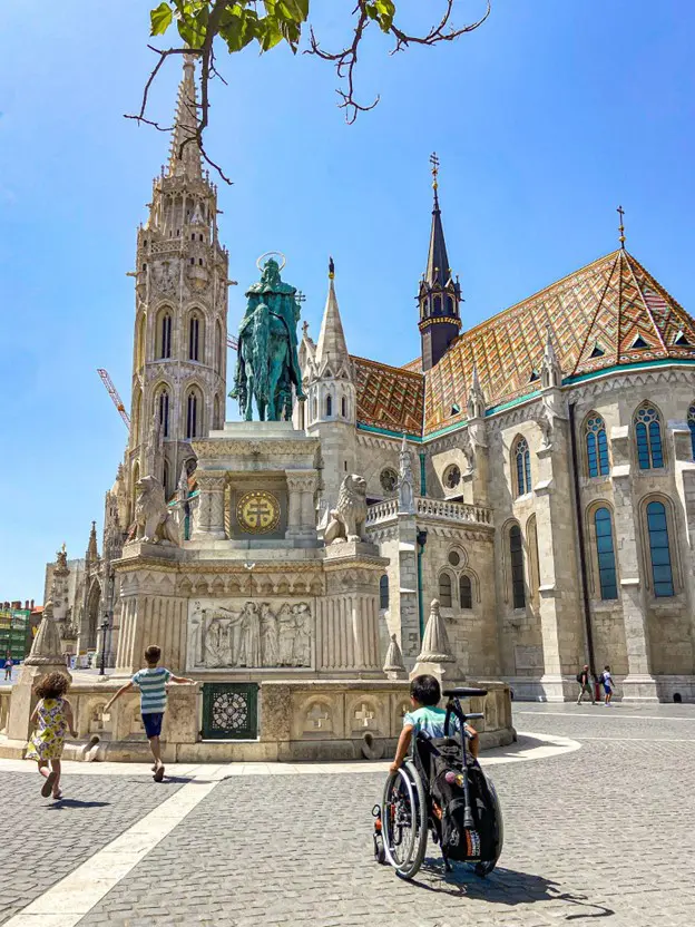 A child in a wheelchair near a historic statue and beautiful buildings under a bright blue sky, while people stroll around.