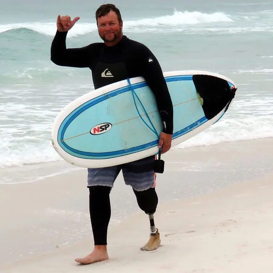 A man in a wetsuit walks on the beach, holding a surfboard and making a shaka sign with his hand, with waves in the background.