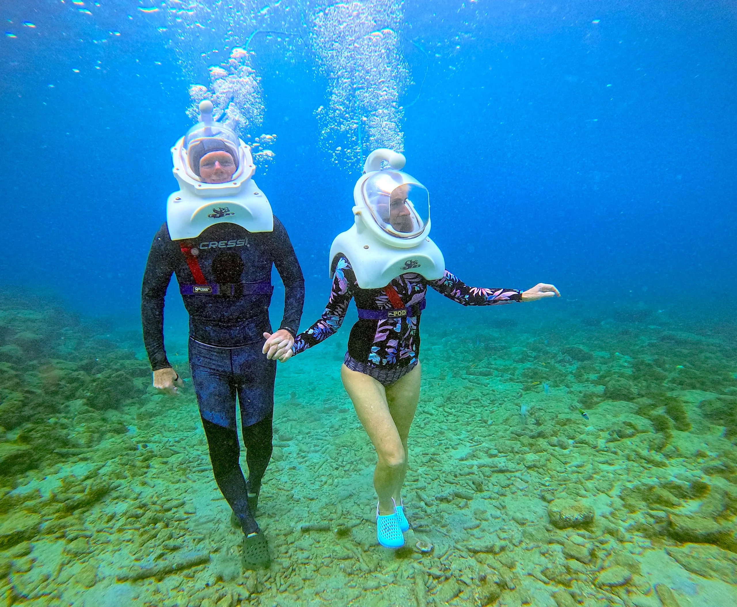 Two divers explore an underwater scene, wearing helmet diving gear, while holding hands amidst bubbles and rocky ocean floor.