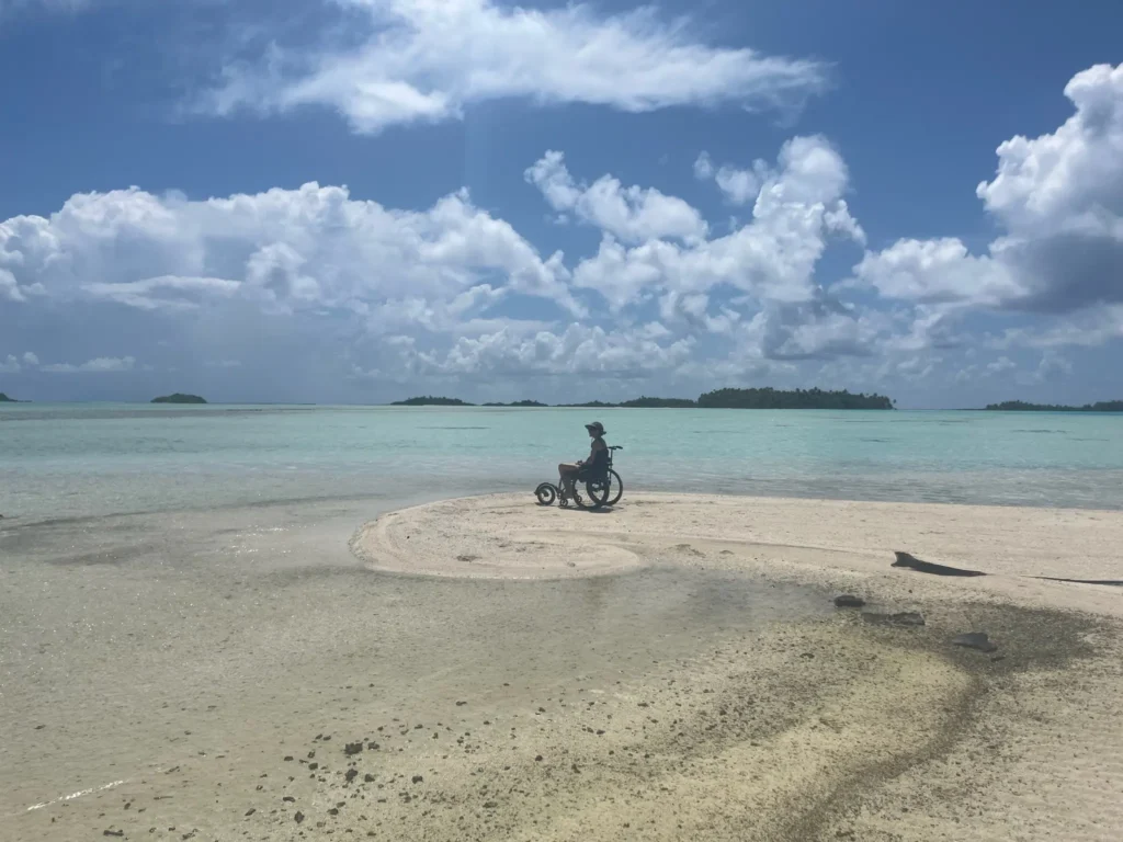 A person on a wheelchair sits on a sandy beach, surrounded by turquoise waters and scattered islands under a blue sky with fluffy clouds.