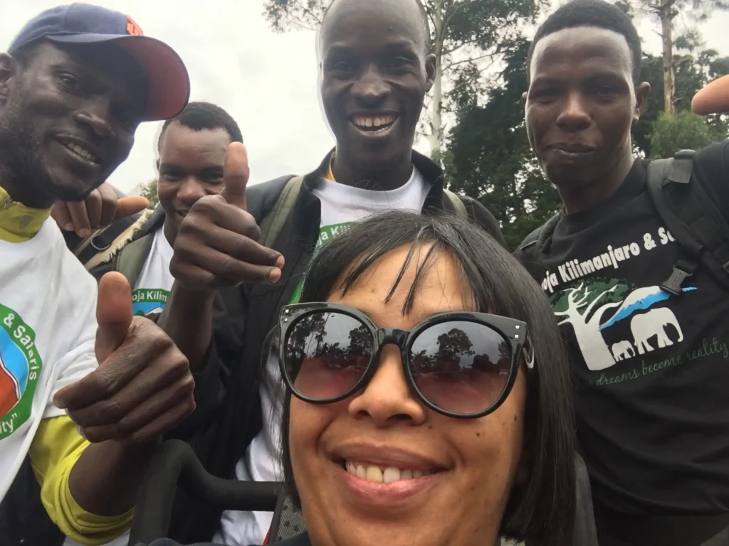 A group of five individuals wearing matching t-shirts, smiling and posing for a photo outdoors among trees.