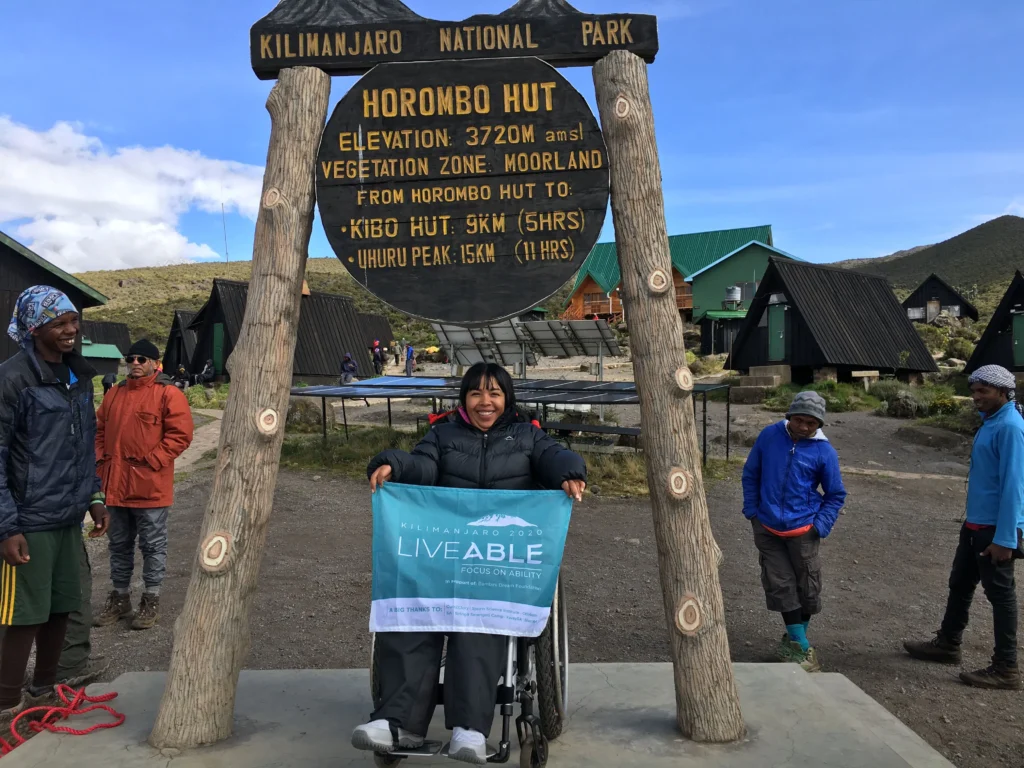 A person in a wheelchair holds a banner at the Horombo Hut sign, surrounded by hikers and mountain lodges at Kilimanjaro National Park.