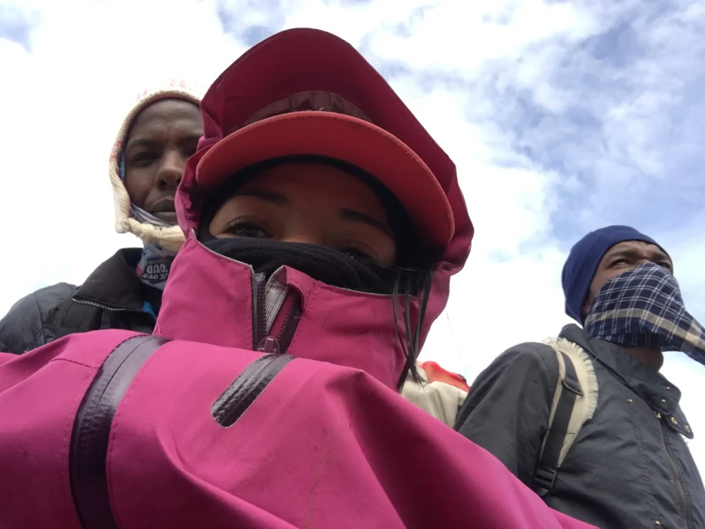 Three individuals wearing winter clothing, including a pink jacket and a red hat, stand against a backdrop of cloudy skies.