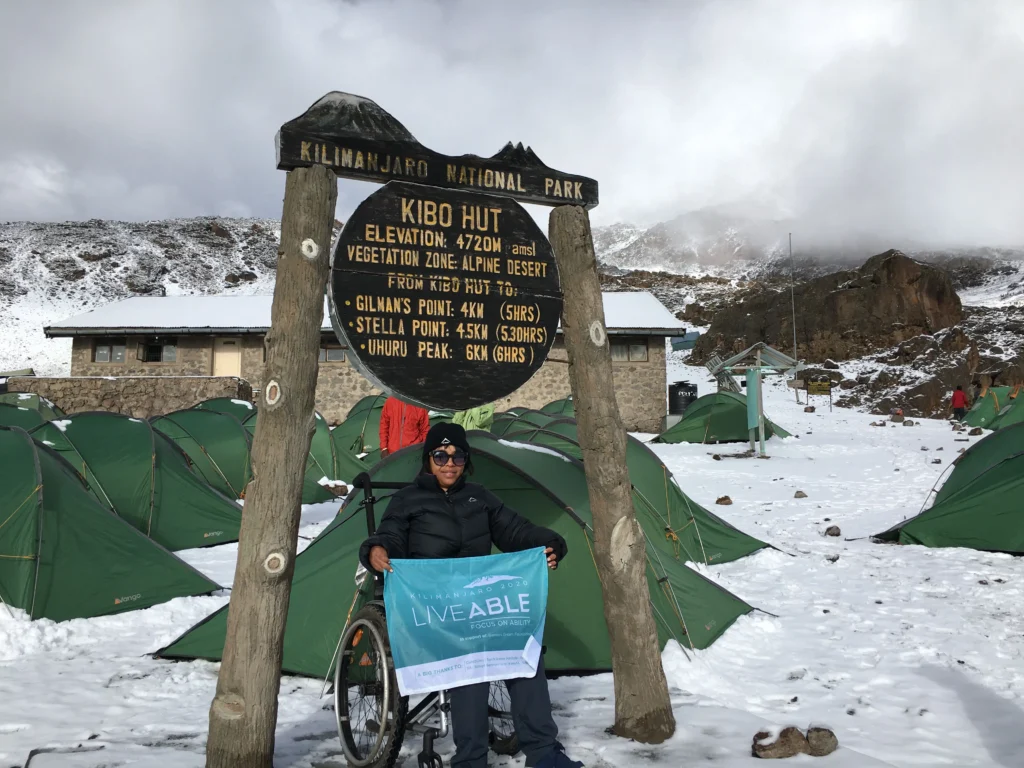 A person in a wheelchair sits beside a sign for Kibo Hut in snowy Kilimanjaro National Park, surrounded by green tents.