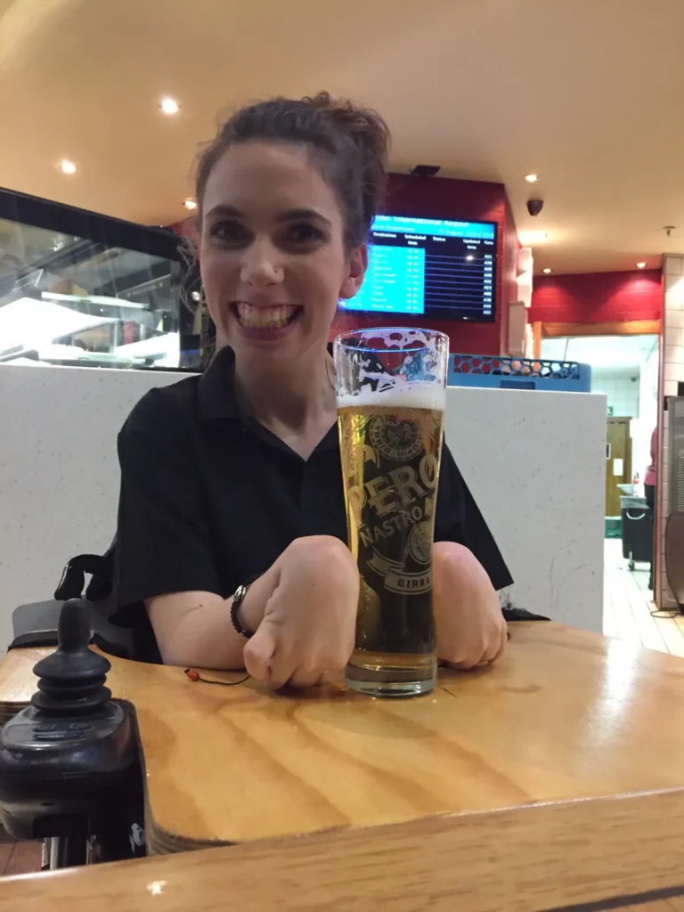 A glass of beer sits on a wooden table, accompanied by hands resting on the surface, in a casual dining area.