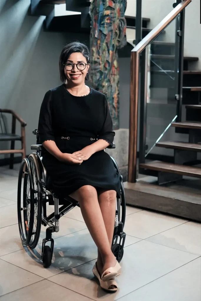 A woman in a black dress sits in a wheelchair, displaying a serene expression in a well-lit indoor setting with stylish decor.
