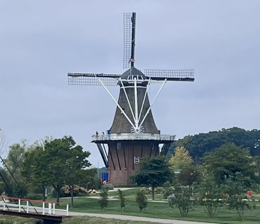 A large, traditional windmill stands amidst lush greenery and cloudy skies. The scene conveys a peaceful, pastoral feel with a sense of heritage.