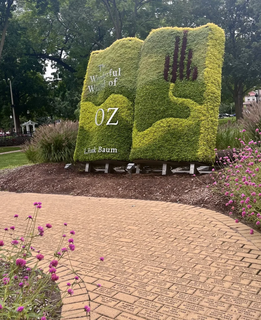 A large book-shaped hedge sculpture displays the title "The Wonderful Wizard of Oz" by L. Frank Baum. Nearby, a yellow brick path with engraved names leads through a vibrant garden with purple flowers, under lush green trees.
