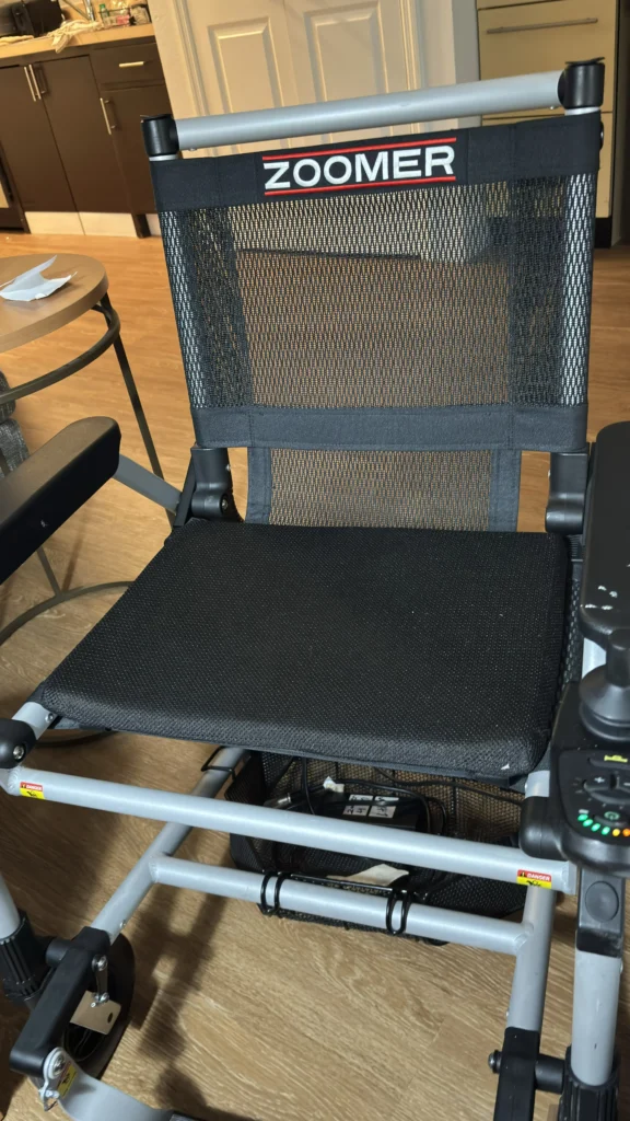 A close-up of a black Zoomer wheelchair seat with a mesh back, located in a well-lit room with wooden flooring and a small table nearby.
