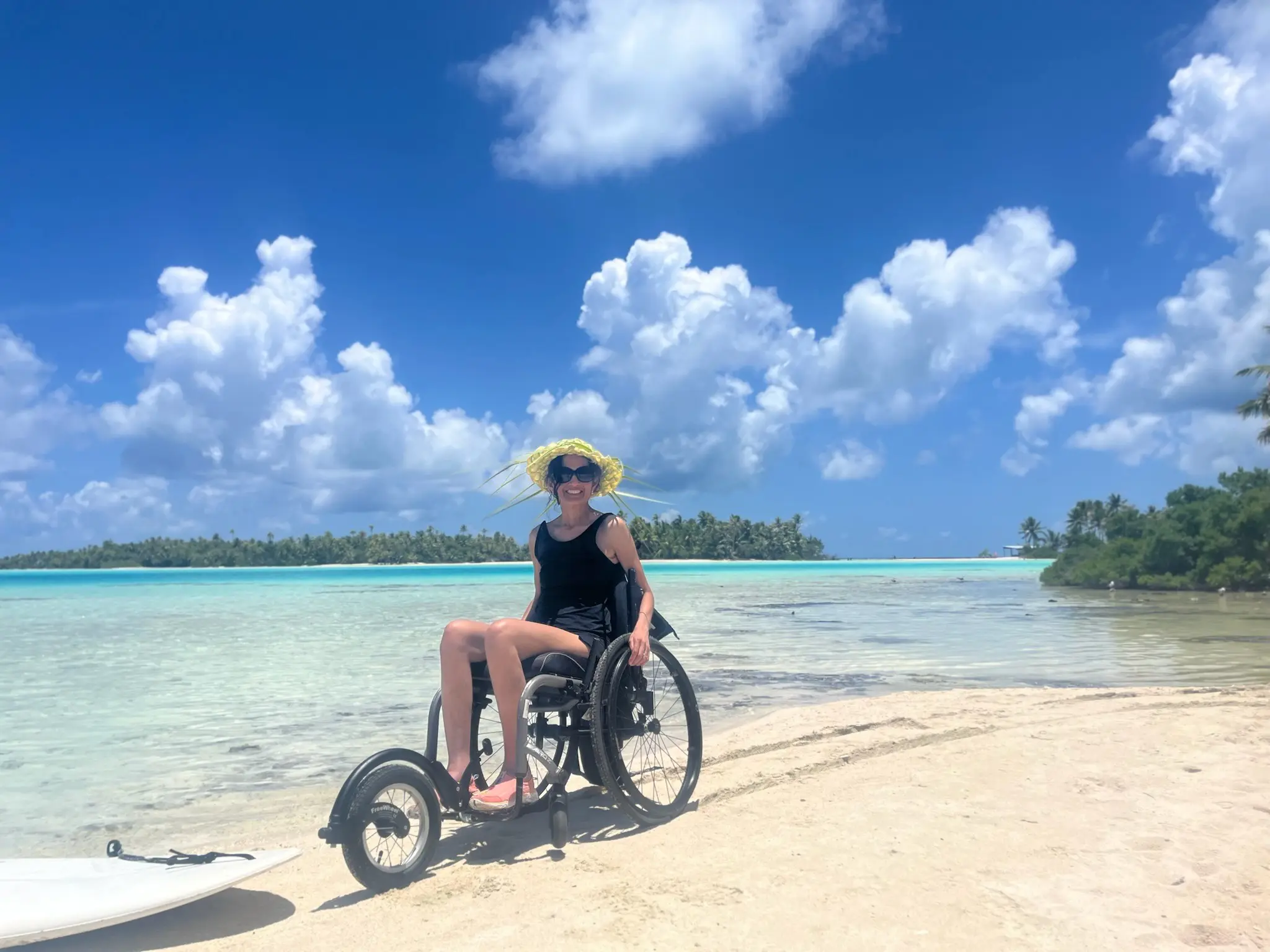 A lady in a wheelchair sits on a sandy beach, with crystal-clear water and lush green trees in the background under a bright blue sky.