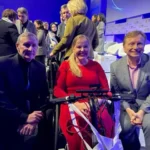 Three individuals pose together at a conference, one seated in a wheelchair, wearing a red dress, surrounded by a bustling audience.