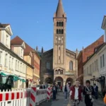 A bustling street with people walking, framed by historic buildings and a tall church tower against a clear blue sky.
