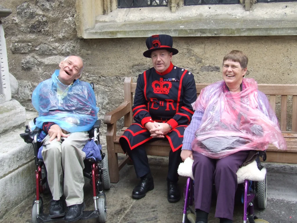 Two individuals in wheelchairs wearing plastic ponchos sit on a wooden bench beside a man dressed in a traditional Yeoman Warder uniform.