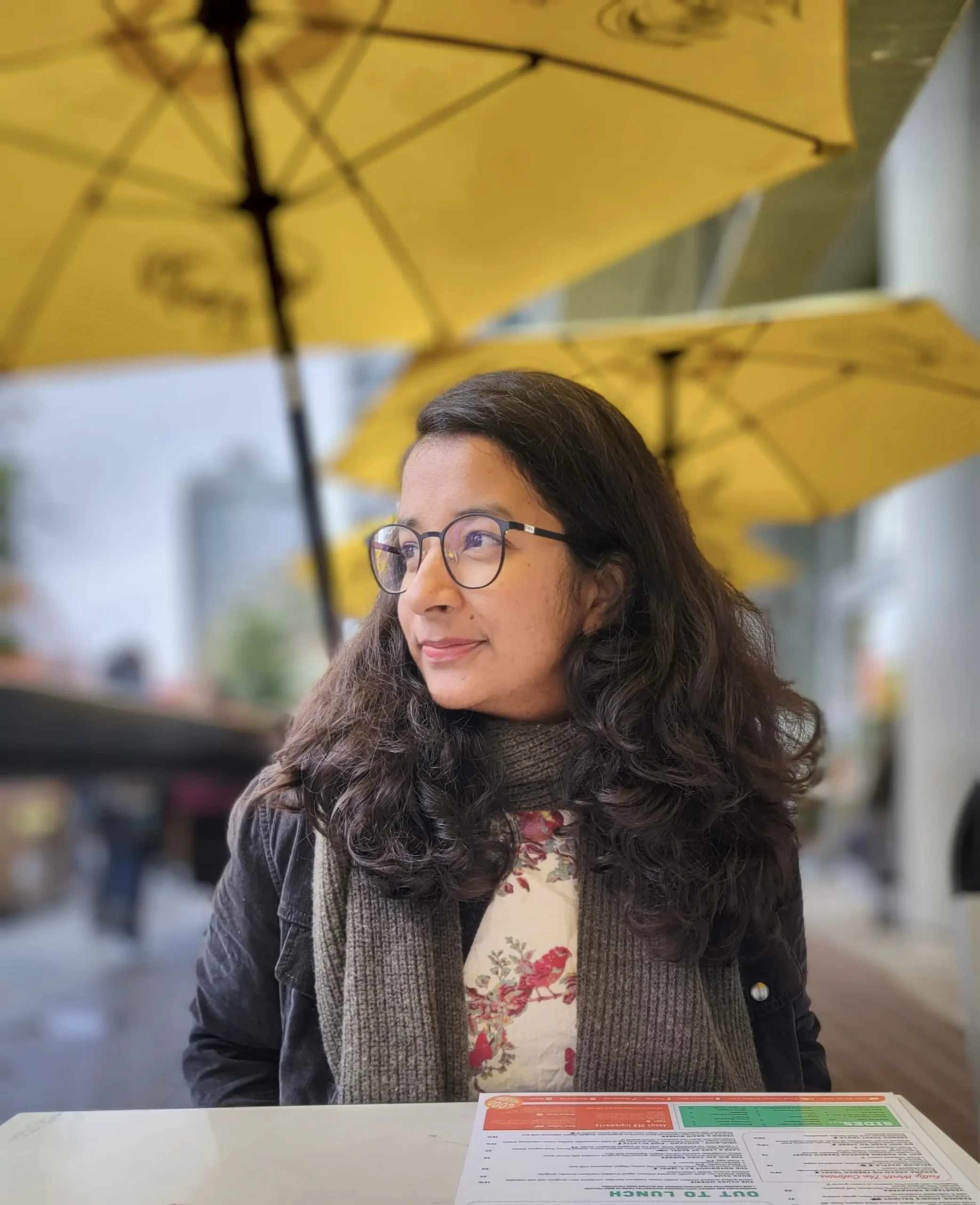 A person with long, wavy hair and glasses sits at a table under a yellow umbrella, with a menu in front of them.