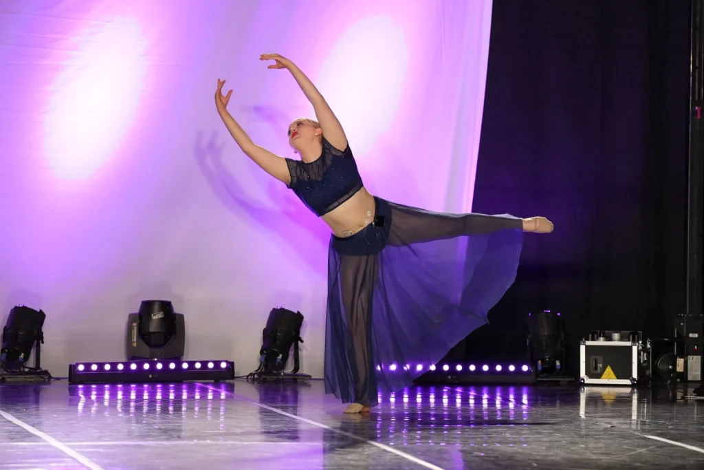 A dancer in a flowing navy costume performs an elegant pose on stage, illuminated by soft purple lighting and surrounded by spotlights.