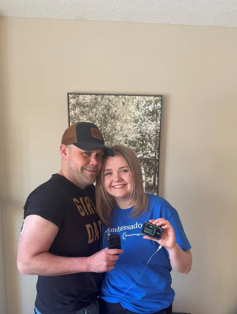 A woman and a man standing together in a warmly lit room, one holding a device, while the other displays a small electronic monitor.