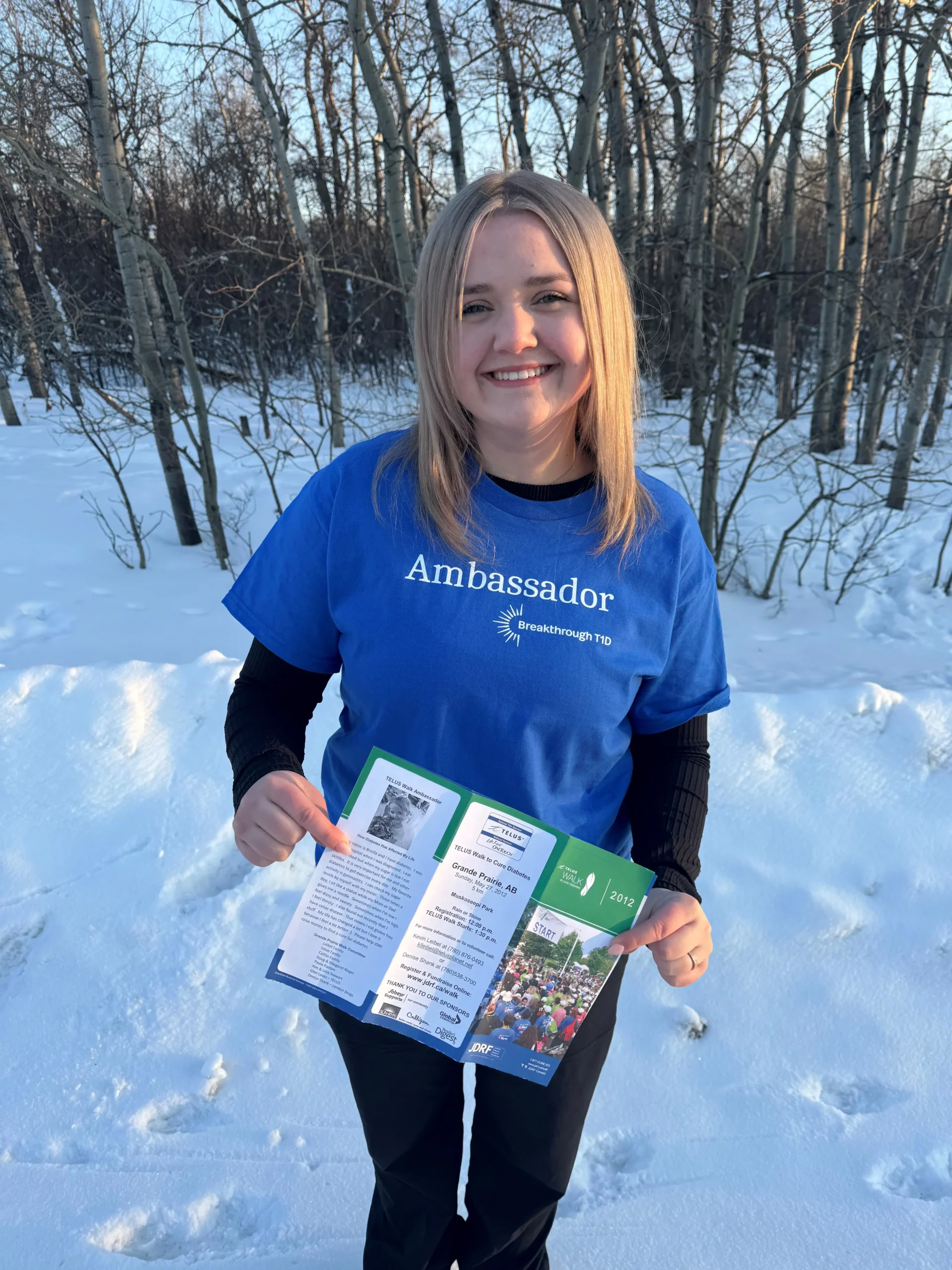 A person in a blue "Ambassador" shirt stands in a snowy forest, holding an information brochure about a community event.