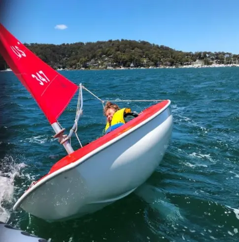 A child in a yellow life jacket leans precariously in a small sailboat with a red sail, navigating choppy waters on a sunny day.