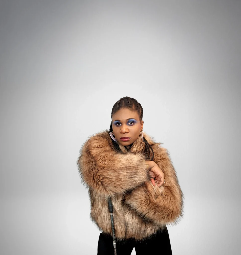 A person in a stylish brown fur coat strikes a pose against a soft gray background, showcasing confidence and individuality.