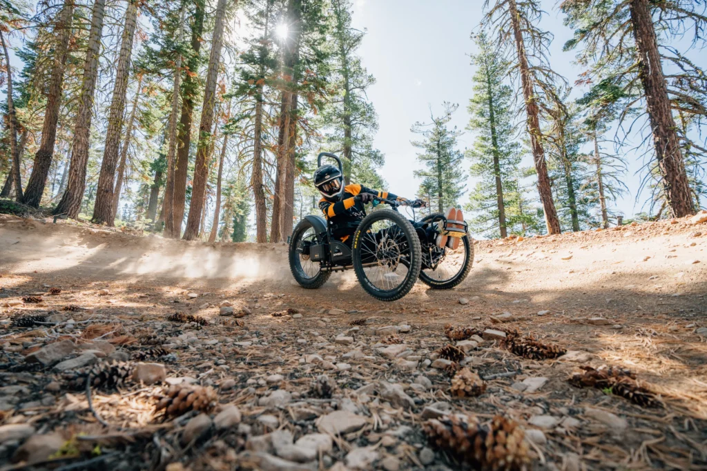 A rider in a helmet speeds around a dirt curve on an off-road handcycle, surrounded by tall trees and dust clouds.