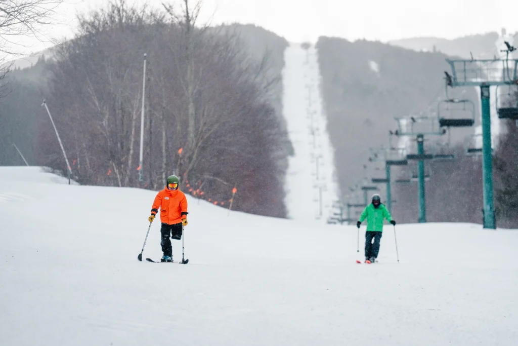 Two skiers traverse a snowy slope in bright orange and green attire, with trees and a ski lift visible in the background.