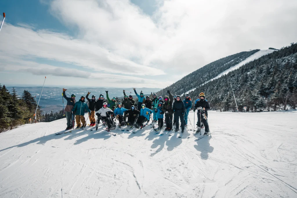 A lively group of skiers poses on a snowy mountain slope, surrounded by pine trees and distant hills under a partly cloudy sky.