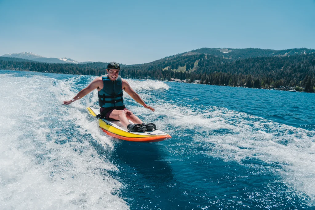 A person in a life jacket rides a colorful surfboard on a vibrant blue lake, with mountains and trees in the background.